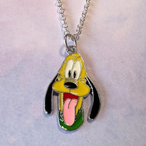 May include: A silver chain necklace with a pendant featuring a yellow cartoon dog with black ears, a pink tongue, and a green collar. The dog's face is white with black eyes and a black nose.