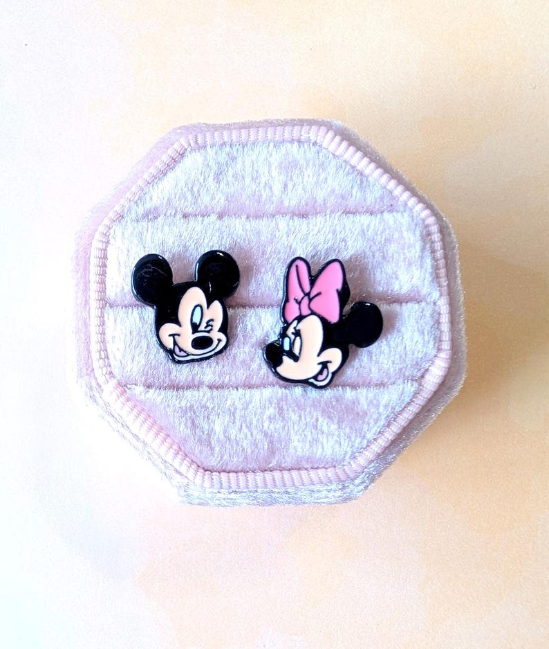 Puede incluir: Un par de pendientes de bot&oacute;n de Mickey Mouse y Minnie Mouse en blanco y negro con detalles en rosa. Los pendientes se muestran en una caja de joyer&iacute;a de terciopelo rosa claro con forma octogonal.