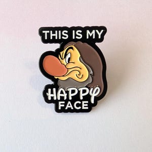 May include: A black and white pin featuring a cartoon character with a large red nose and a grumpy expression. The text "THIS IS MY HAPPY FACE" is above and below the character's face.