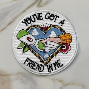 May include: Embroidered patch with the text "YOU'VE GOT A FRIEND IN ME". The patch features a heart-shaped design with a handshake, a green and white space ship, and a cowboy-style sleeve. The patch is round with a white border.