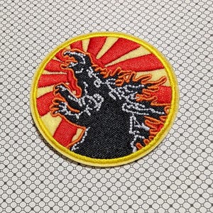 May include: Embroidered patch featuring a black Godzilla silhouette against a red and yellow sunburst design. The patch has a yellow border and is likely for application on clothing or accessories.