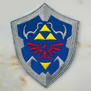 May include: A blue and silver embroidered patch featuring the iconic Triforce symbol from the Legend of Zelda video game series. The Triforce is surrounded by a stylized design of a shield.