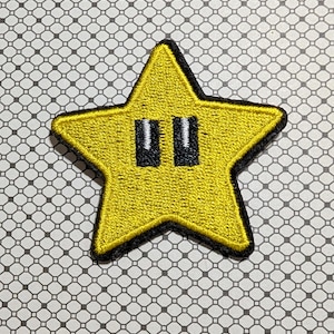 May include: Embroidered yellow star patch with black outline and two black rectangles representing eyes. The patch is on a patterned background.