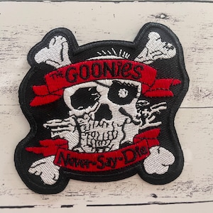 May include: Black and white embroidered patch with a skull and crossbones. The skull is wearing an eye patch and has a red bandana with the text "The Goonies" on it. The bandana has the text "Never Say Die!" on it.