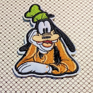 May include: Embroidered patch featuring Goofy from Disney. The patch shows Goofy in his classic pose, with a green hat, orange jacket, and black ears. The patch is outlined in black and white.