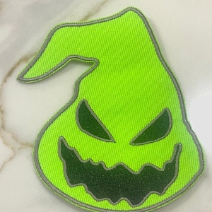 May include: A bright green felt patch with a stitched outline, depicting a cartoon ghost with a wide, toothy grin and large eyes.