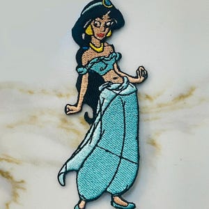 May include: An embroidered iron-on patch featuring a cartoon image of Princess Jasmine from Disney's Aladdin. She is wearing a blue outfit with a gold belt and a blue head scarf.