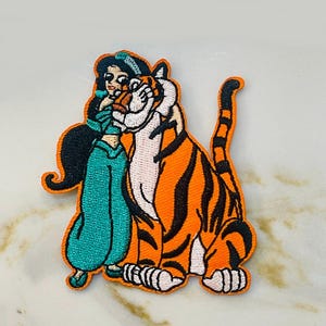 May include: Embroidered patch featuring a cartoon depiction of a woman in teal clothing embracing a large orange tiger with black stripes. The patch has a white border and is likely for clothing or accessories.