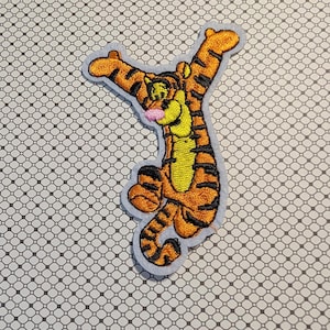 May include: Embroidered patch featuring Tigger from Winnie the Pooh. The patch shows Tigger in a jumping pose with orange and black stripes, yellow belly, and a pink nose. The patch has a light blue border.