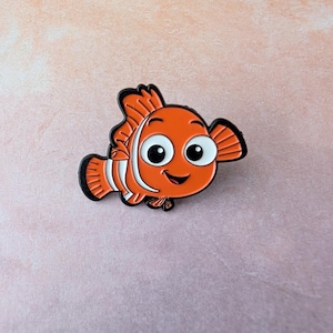 May include: Orange and white cartoon fish pin with black outlines. The fish has large, expressive eyes and a smiling mouth. The pin is likely made of enamel or a similar material, and is set against a pink background.