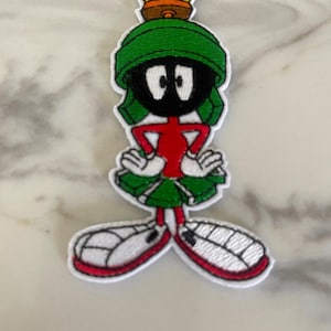 May include: A green, red, and black embroidered iron-on patch of the cartoon character Marvin the Martian from the Looney Tunes series. The patch features Marvin's signature helmet, red suit, and large black eyes.