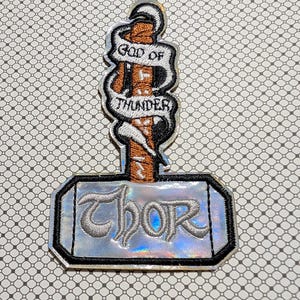 May include: Embroidered patch in the shape of Thor's hammer, with the word "Thor" in silver script on the hammer head. The handle features the words "God of Thunder" on a banner.