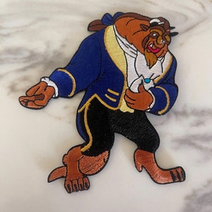 May include: An embroidered iron-on patch featuring a cartoon image of the Beast from Disney's Beauty and the Beast. The Beast is wearing a blue jacket with gold trim, a white shirt, and black pants. He is holding a blue rose in his right hand.