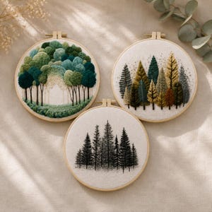 Pode incluir: Três bastidores de bordado circulares de madeira com arte em tecido. O bastidor superior esquerdo apresenta uma floresta densa de árvores verdes. O superior direito mostra uma variedade de árvores em tons de verde, amarelo e castanho. O inferior mostra uma floresta de pinheiros preta.