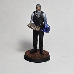 May include: A painted miniature figurine of a man with a white beard wearing a white shirt, a black waistcoat, and black trousers. He is holding a book in his left hand and a blue glowing orb in his right hand. The figurine is standing on a wooden base.