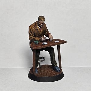 May include: A painted miniature figurine of a person sitting at a table with cards in their hand. The person is wearing a brown jacket and has short brown hair. The table is brown wood and has a black base.
