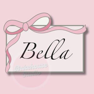 May include: A pale pink background with a rectangular card displaying the name "Bella" in cursive script. A pink ribbon bow decorates the top left corner. The words "Bakehouse Studio" are at the bottom.
