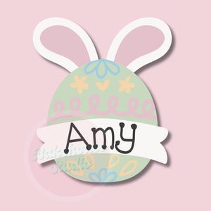 Könnte beinhalten: Ein Osterei-Design mit Hasenohren, mit dem Namen "Amy" auf einem Banner. Das Ei ist hellgrün mit pastellfarbenen Blumen- und Wirbeldekorationen. Der Hintergrund ist ein zartes Rosa.