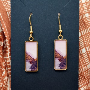 May include: A pair of gold dangle earrings with rectangular resin charms featuring a watercolor landscape scene. The charms are a light pink color with brown and gray mountains and a body of water.