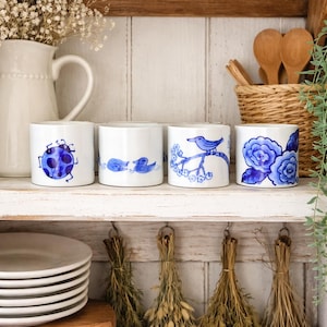May include: Four white ceramic mugs with blue hand-painted designs. The designs include a ladybug, two ducks, a bird on a branch, and blue flowers. The mugs are displayed on a white shelf with other kitchen items.