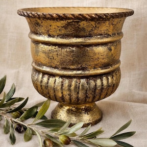 May include: A gold-colored decorative urn with a textured finish. The urn has a wide, fluted base and a detailed rim. Olive branches with green leaves and olives are in the foreground, adding a natural element to the composition. The urn is likely made of metal or a similar material.