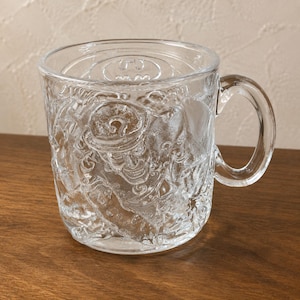 May include: A clear glass mug featuring a raised, embossed design of the Riddler character wearing a bowler hat with a question mark, along with a Batman logo at the top. The mug has a smooth interior, a sturdy handle, and a textured exterior surface.