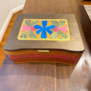 May include: A rectangular wooden box with a dark brown lid featuring a hand-painted design of a central blue flower flanked by green leaves and pink petals. The base of the box consists of stacked layers in shades of orange, red, and yellow.