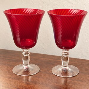 May include: A pair of matching wine glasses featuring deep red bowls with a subtle, swirling ribbed texture. Each glass is supported by a clear, transparent glass stem and a circular base, providing a stable foundation for the vibrant red vessels.