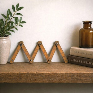 May include: A vintage wooden folding ruler, with visible measurement markings, is displayed open on a wooden shelf. The ruler is a warm, aged yellow color. A small brown glass bottle and two books are in the background. A small plant in a beige pot is on the left.