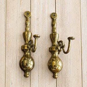 May include: Two antique brass wall sconces with a decorative design. Each sconce features a bulbous base, a curved arm, and a top with a hole for mounting. The sconces are mounted on a light-colored wooden surface.