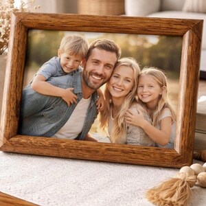 May include: A rectangular wooden picture frame with a natural grain finish containing a photograph of a family. The photo shows a smiling adult with a young child on their back, next to another adult and a young child, all outdoors in sunlight.