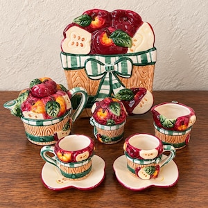 May include: A ceramic tea set featuring a teapot, creamer, two cups with saucers, and a large serving tray. Each piece is decorated with a motif of red apples, green leaves, and a green and white checkered pattern over a wood-grain texture.