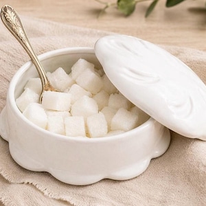 May include: A white ceramic sugar bowl filled with sugar cubes, with a decorative lid and a gold spoon inside. The bowl has a scalloped edge and sits on a neutral-colored cloth.
