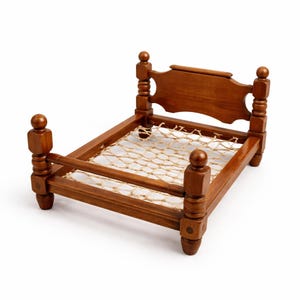 May include: A miniature wooden bed with a dark brown finish. The bed frame has rounded posts and a headboard with a decorative cutout. The bed's base is made of woven natural-colored rope, creating a rustic aesthetic. This small bed is likely for dolls or pets.