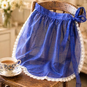 May include: A sheer blue apron with a white lace trim and a matching bow. The apron is draped over a wooden chair, with a teacup and saucer in the foreground. The teacup has a floral design.