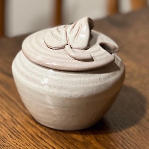 May include: A small, round, off-white ceramic jar with a speckled finish and a matching lid. The lid features a sculpted, raised leaf design and a small notch on the side for a spoon. The body of the jar has subtle horizontal ridges.