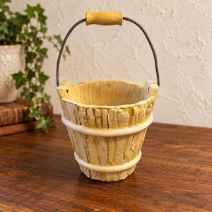 May include: A small, cream-colored bucket with a textured, vertical slat design and two horizontal bands. It features a dark metal wire handle with a yellow wooden grip at the top, designed for holding small items or decorative displays.