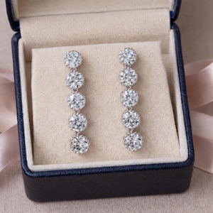 May include: A pair of elegant drop earrings featuring a series of sparkling, round-cut gemstones. The earrings are displayed inside a blue jewelry box with a cream-colored interior, and a pink ribbon is visible in the background.