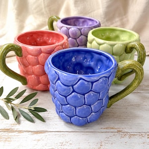 May include: Four ceramic mugs in shades of pink, purple, green, and blue. Each mug has a textured, scale-like pattern and a green handle. The mugs are arranged on a white wooden surface.