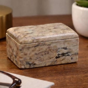 May include: A rectangular marble box with a lid. The box is a mix of beige, brown, and gray tones, with darker veining patterns. The box is sitting on a wooden surface.