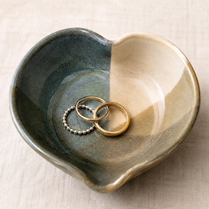 May include: A heart-shaped ceramic dish featuring a two-tone glaze with one side in deep blue-grey and the other in a creamy beige. Two gold-toned metal rings are resting inside the bowl, one plain band and one with a beaded texture.