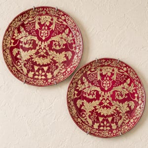 May include: Two circular ceramic plates featuring a deep red background with intricate gold-colored floral and acanthus leaf patterns. Each plate is held by silver-toned metal wall mounting clips, displaying a symmetrical design across the surface.
