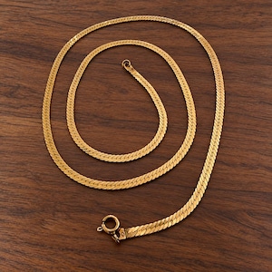 May include: A gold-toned herringbone chain necklace featuring a flat, flexible link design. The necklace is arranged in a circular loop and includes a round spring ring clasp at one end, showcasing a polished, reflective metallic finish.