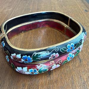 May include: A square-shaped hinged bangle bracelet featuring a black and deep red enamel finish with gold-tone metal edges. The surface is decorated with a floral pattern in blue, white, and green, with a smooth interior and a secure metal clasp.