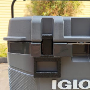 May include: A gray Igloo cooler featuring a black handle and a black textured latch mechanism on the front. The cooler has a diamond-patterned side texture and the white letters IGLO are visible on the front surface of the gray plastic body.