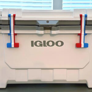 May include: A white Igloo cooler featuring a gray lid and two latch mechanisms on the front. Each latch is accented with vertical stripes in red, white, and blue. The brand name IGLOO is printed in gray capital letters on the center of the cooler.