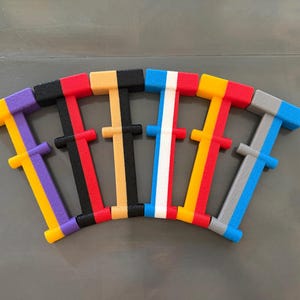 May include: A set of six T-shaped plastic tools arranged in a fan pattern. Each tool features a dual-color design with a textured surface. The color combinations include purple and yellow, black and red, tan and black, blue and white, yellow and red, and gray and blue.