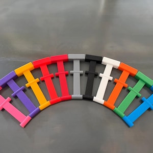 May include: A collection of eleven T-shaped plastic connectors arranged in a semi-circular fan pattern. The connectors are solid colors including pink, purple, yellow, red, gray, black, white, orange, green, and blue, each with a crossbar.