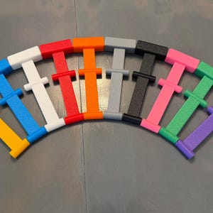 May include: A collection of ten H-shaped plastic connectors arranged in a semi-circular arc. The connectors are solid colors including yellow, blue, white, red, orange, gray, black, pink, green, and purple, each featuring a central crossbar.
