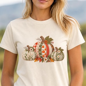 Pode incluir: Uma t-shirt de manga curta na cor creme com um gráfico central de três abóboras. As abóboras são decoradas com vários padrões, incluindo xadrez, bolinhas e motivos florais em tons de laranja, verde e branco com folhas.